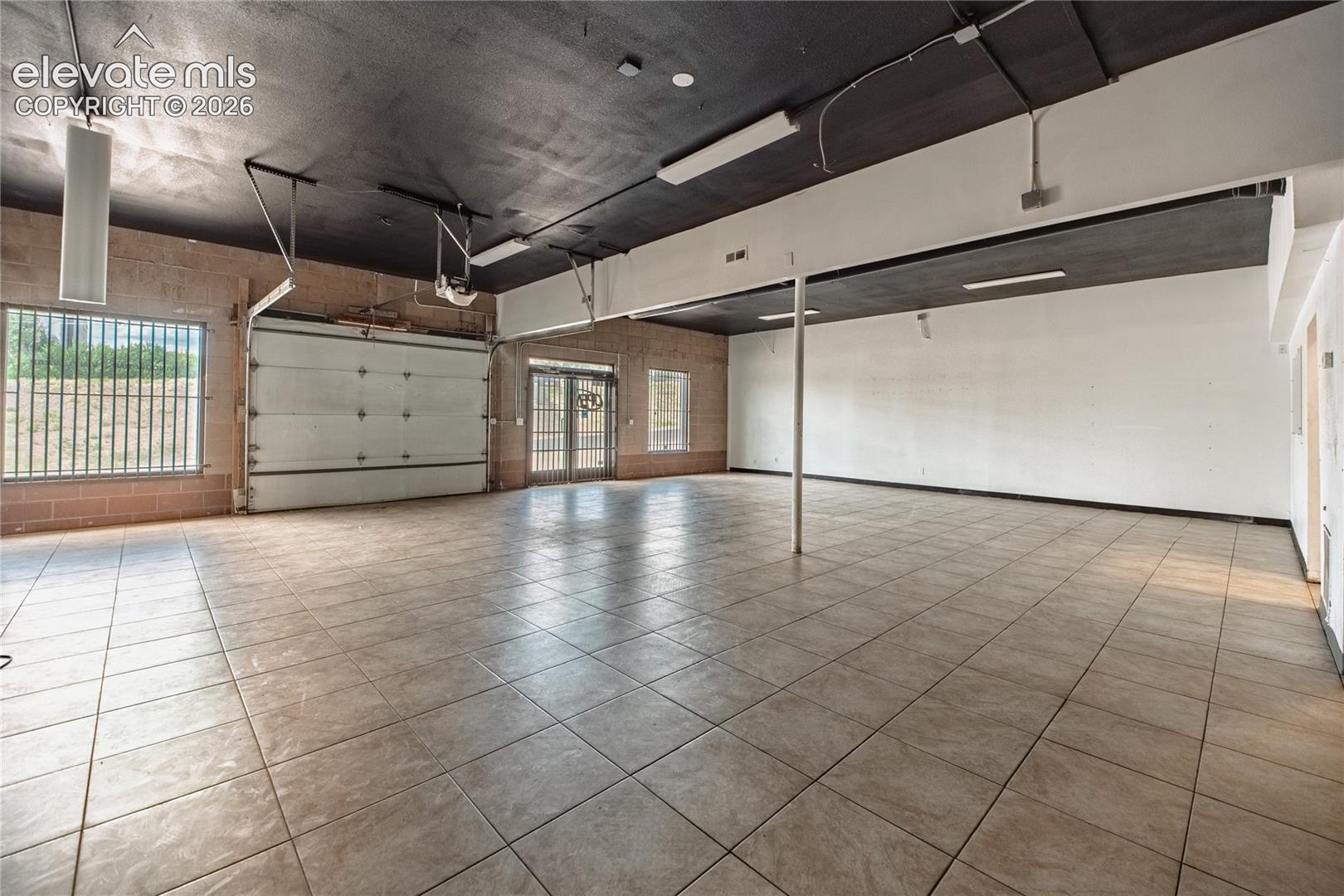 Spacious interior featuring a roll-up garage door, tile flooring, exposed brick walls, and dual entry doors with security bars