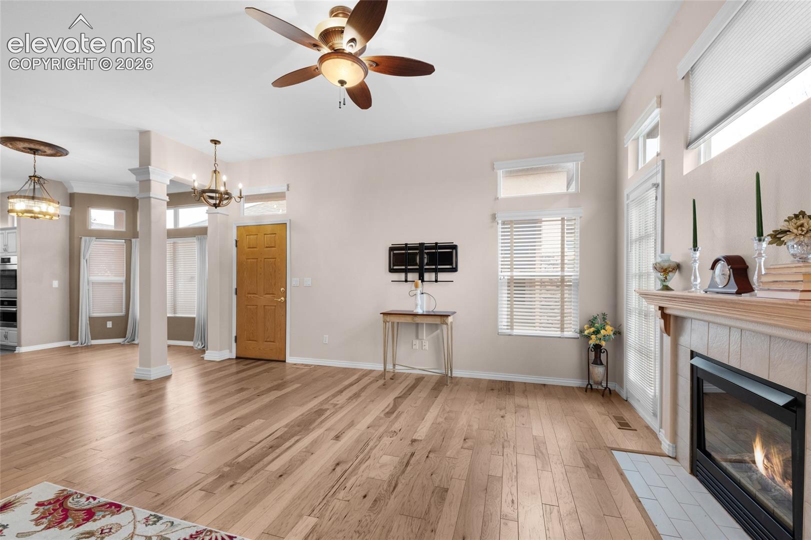 Open-concept interior featuring wood-finish flooring, a fireplace with a light-tone mantel, architectural columns, transom windows, and dual ceiling-mounted light fixtures
