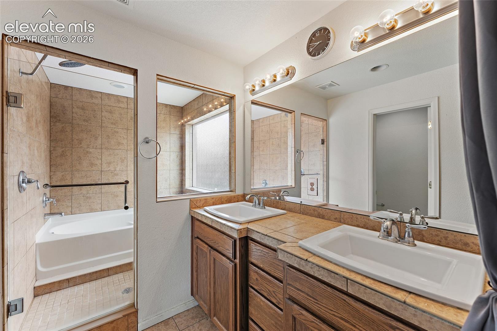 Dual-vanity bathroom featuring tile-surround countertops, a separate soaking tub with tile enclosure, wood cabinetry, and integrated full-width mirrors