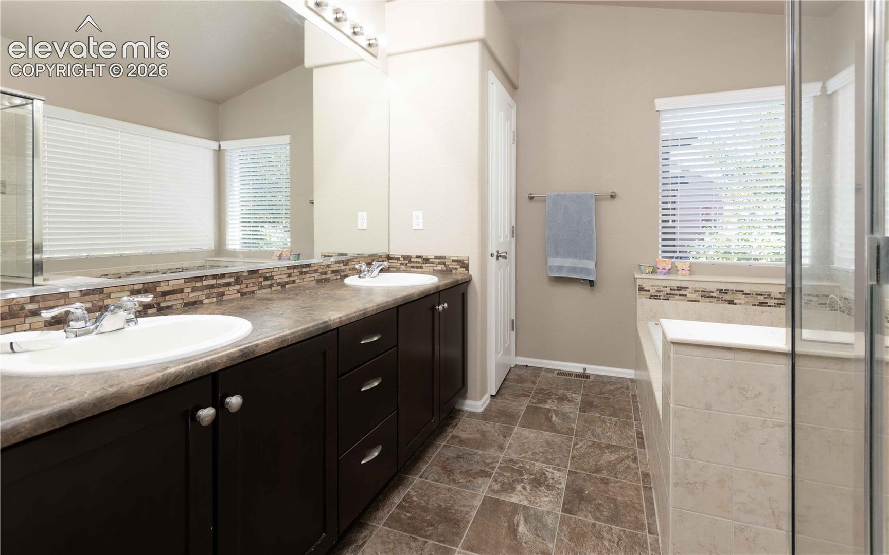 Bathroom featuring a dual-sink vanity with dark cabinetry, stone-finish countertops, tile backsplash, and a glass-enclosed soaking tub