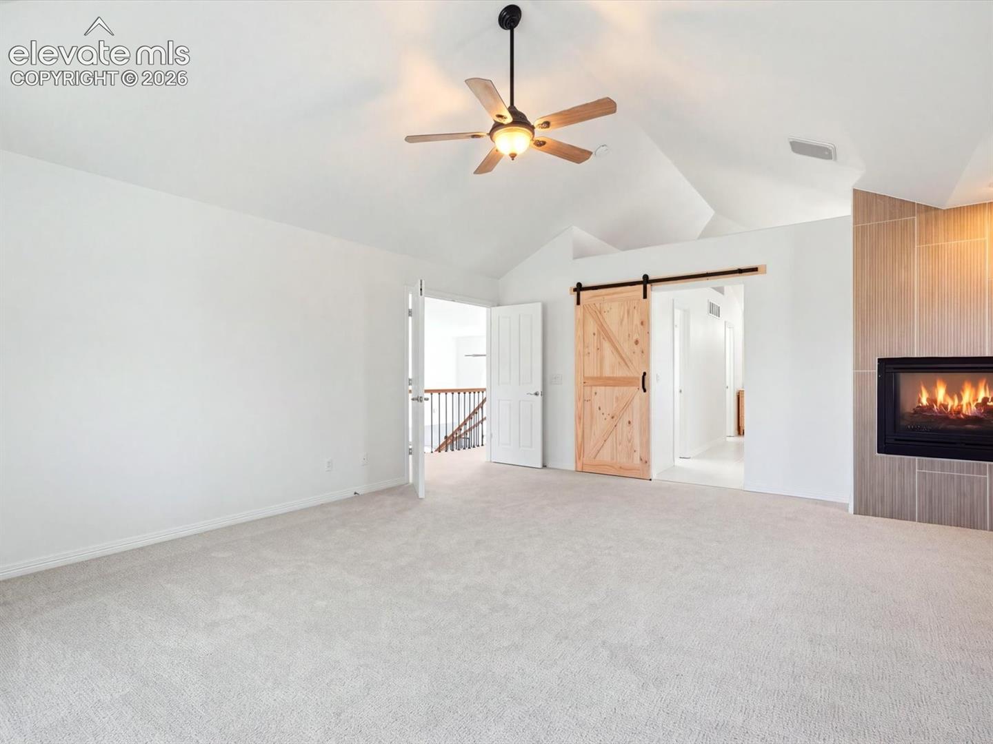Vaulted ceilings, built-in fireplace with neutral tile surround, wooden barn-style sliding door, and ceiling fan with integrated light fixture