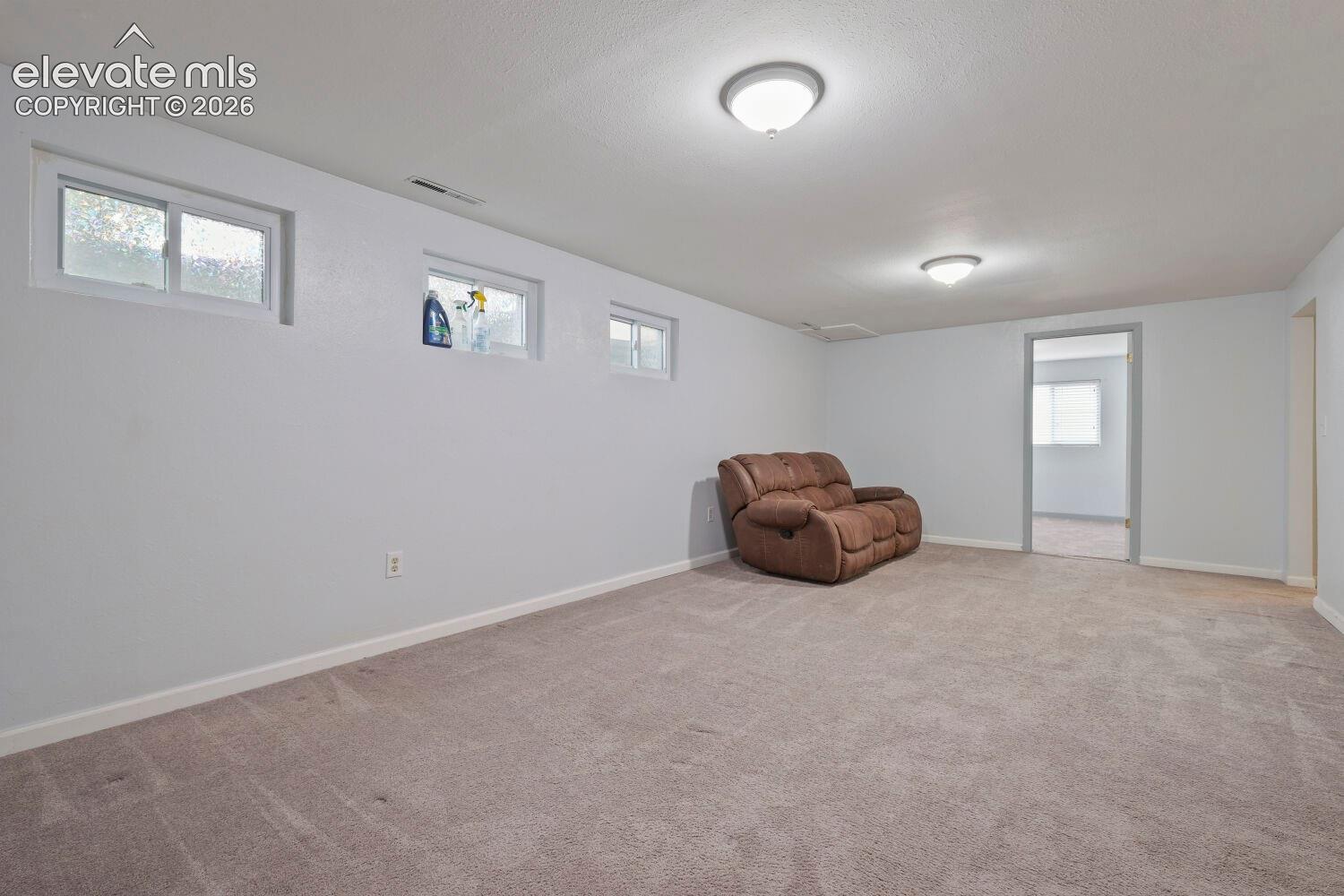 Carpeted room with light gray walls, featuring three high-set windows, two ceiling-mounted dome lights, and a doorway opening into another room