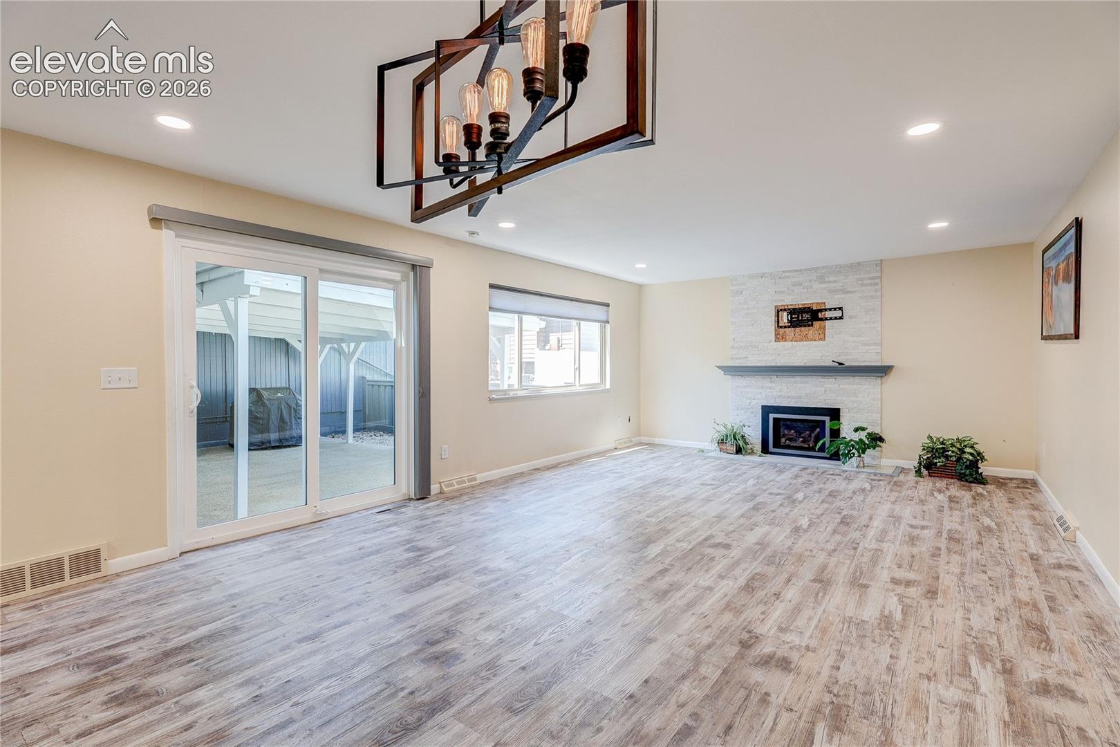 Open-concept living space featuring wood-finish flooring, a stacked-stone fireplace with a mantel, a modern geometric chandelier, recessed lighting, and sliding glass doors