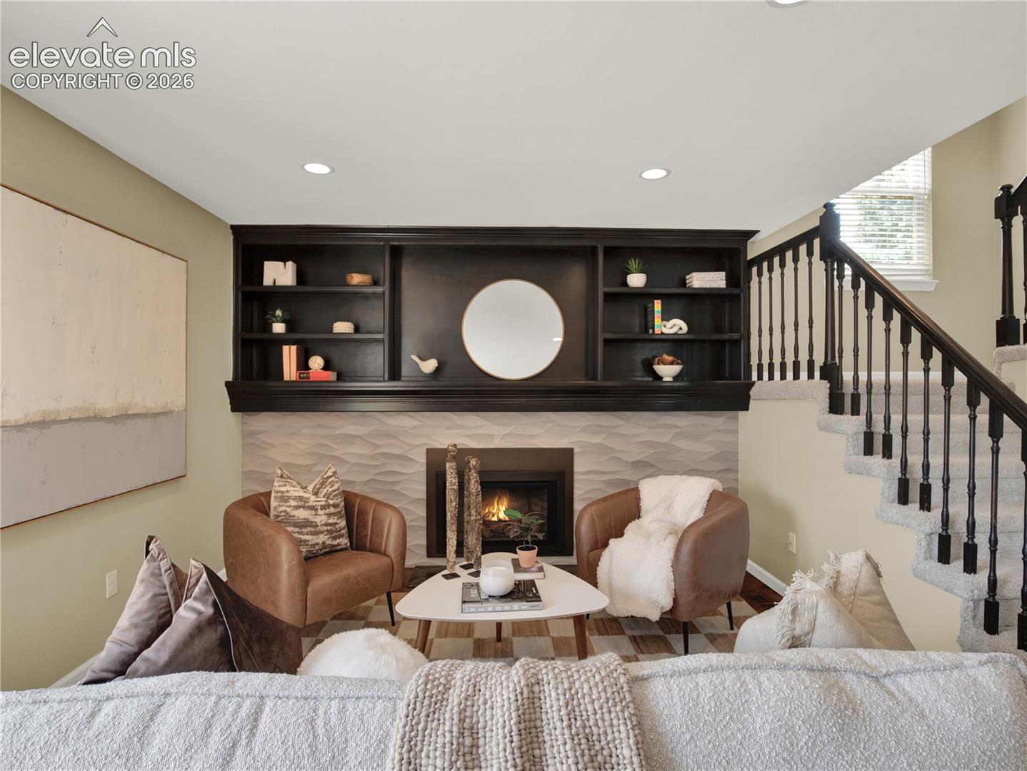 Fireplace feature wall with geometric textured tile surround, black custom built-in shelving, recessed ceiling lighting, and staircase with black balusters