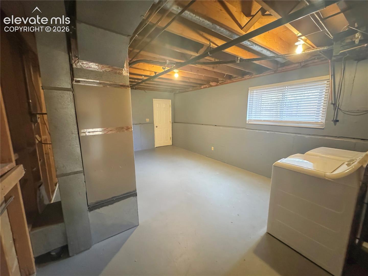Unfinished space featuring painted concrete walls, exposed ceiling joists, metal ductwork, and a horizontal window with blinds