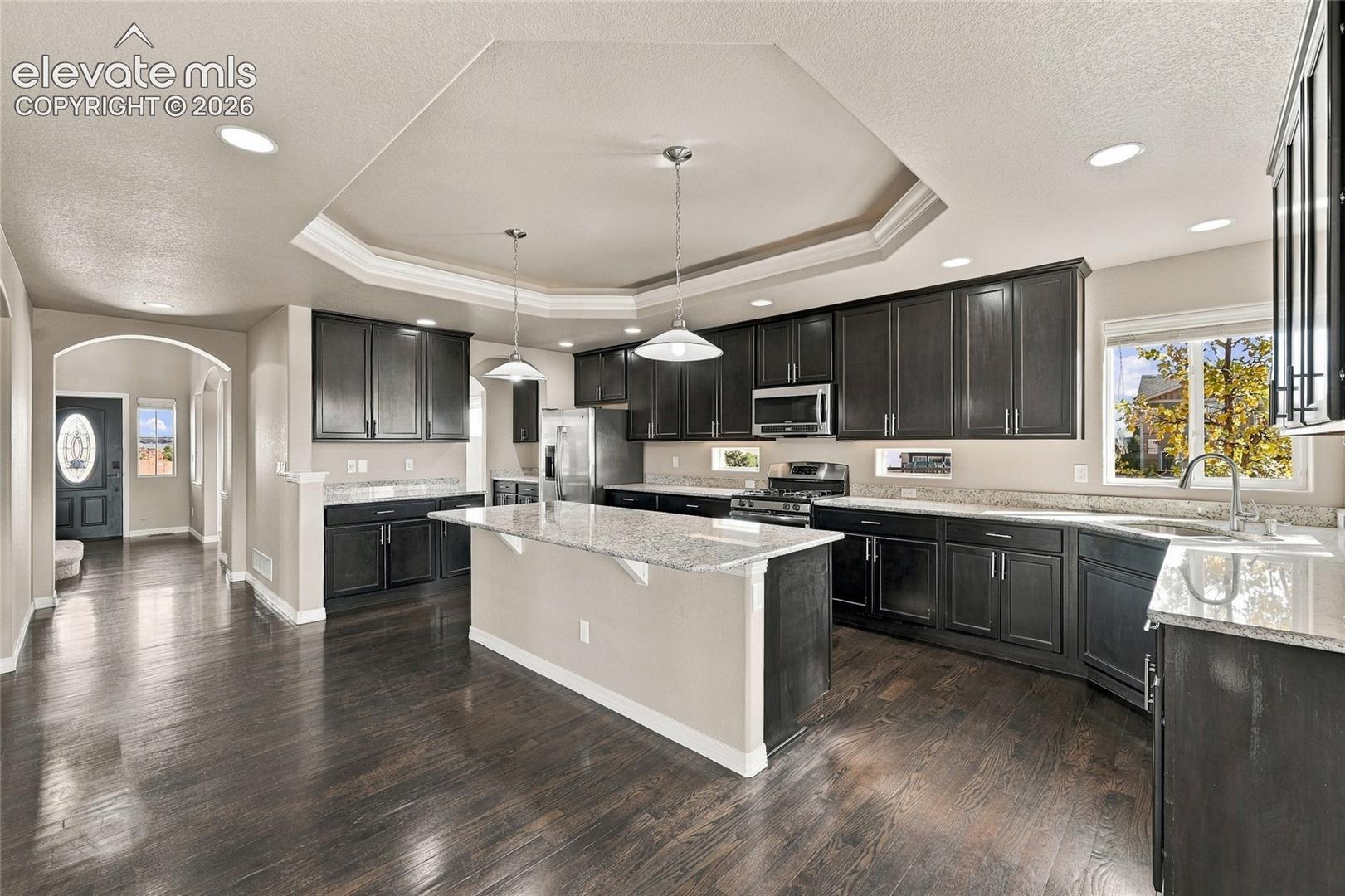 Kitchen featuring dark cabinetry, stone countertops, wood-finish flooring, a central island, and a tray ceiling with pendant lighting