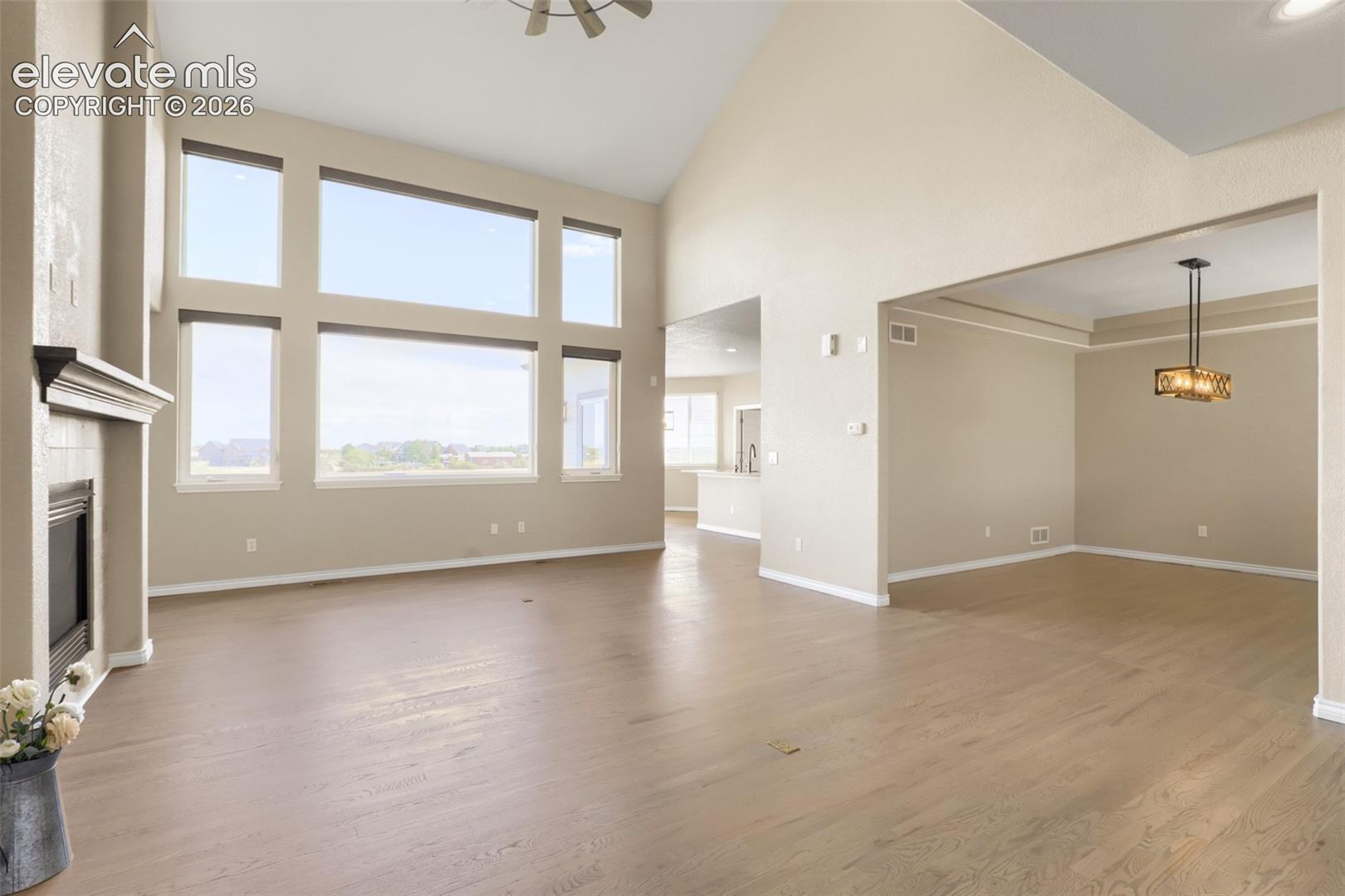 Sunlit, vaulted family room featuring a wall of floor-to-ceiling windows, overhead ceiling fan, and cozy gas fireplace. The expansive open floor plan flows seamlessly into an adjacent formal dining room accented by a tray ceiling and a modern industrial pendant light fixture.
