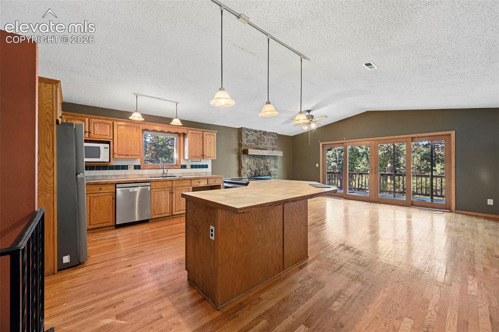 Kitchen and open living area featuring wood-finish flooring, a stone fireplace, wood cabinetry, stainless steel appliances, and a sliding glass door