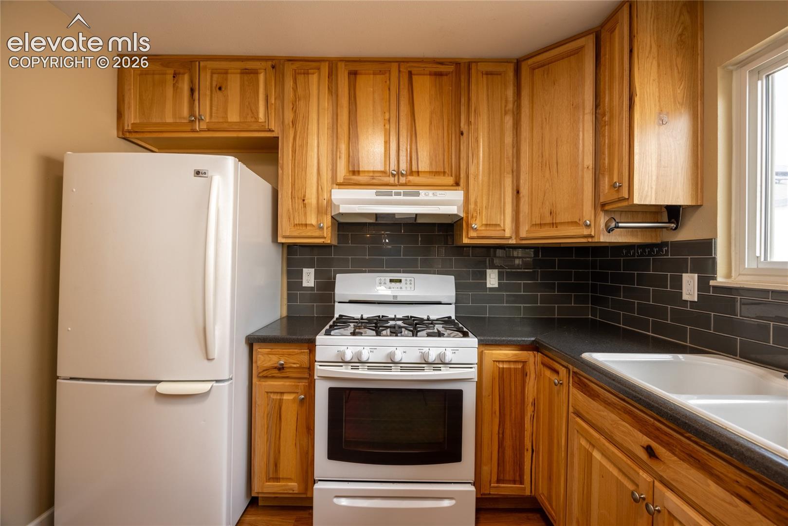 Kitchen featuring wood cabinetry, dark countertops, subway tile backsplash, a white gas range, and a double basin sink