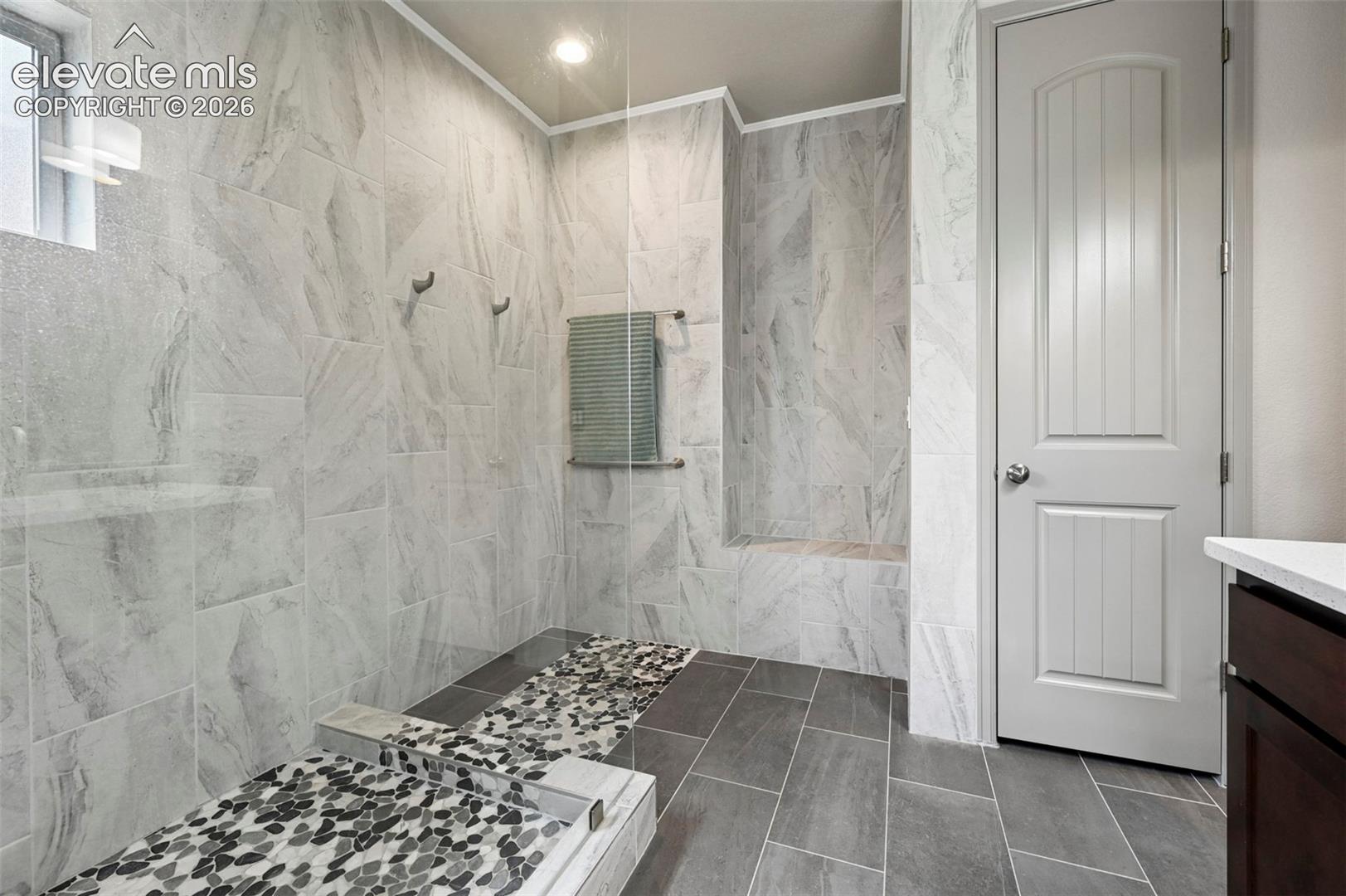Walk-in shower featuring floor-to-ceiling stone-pattern tile, pebble-stone shower floor, built-in tiled bench, frameless glass partition, and recessed ceiling lighting