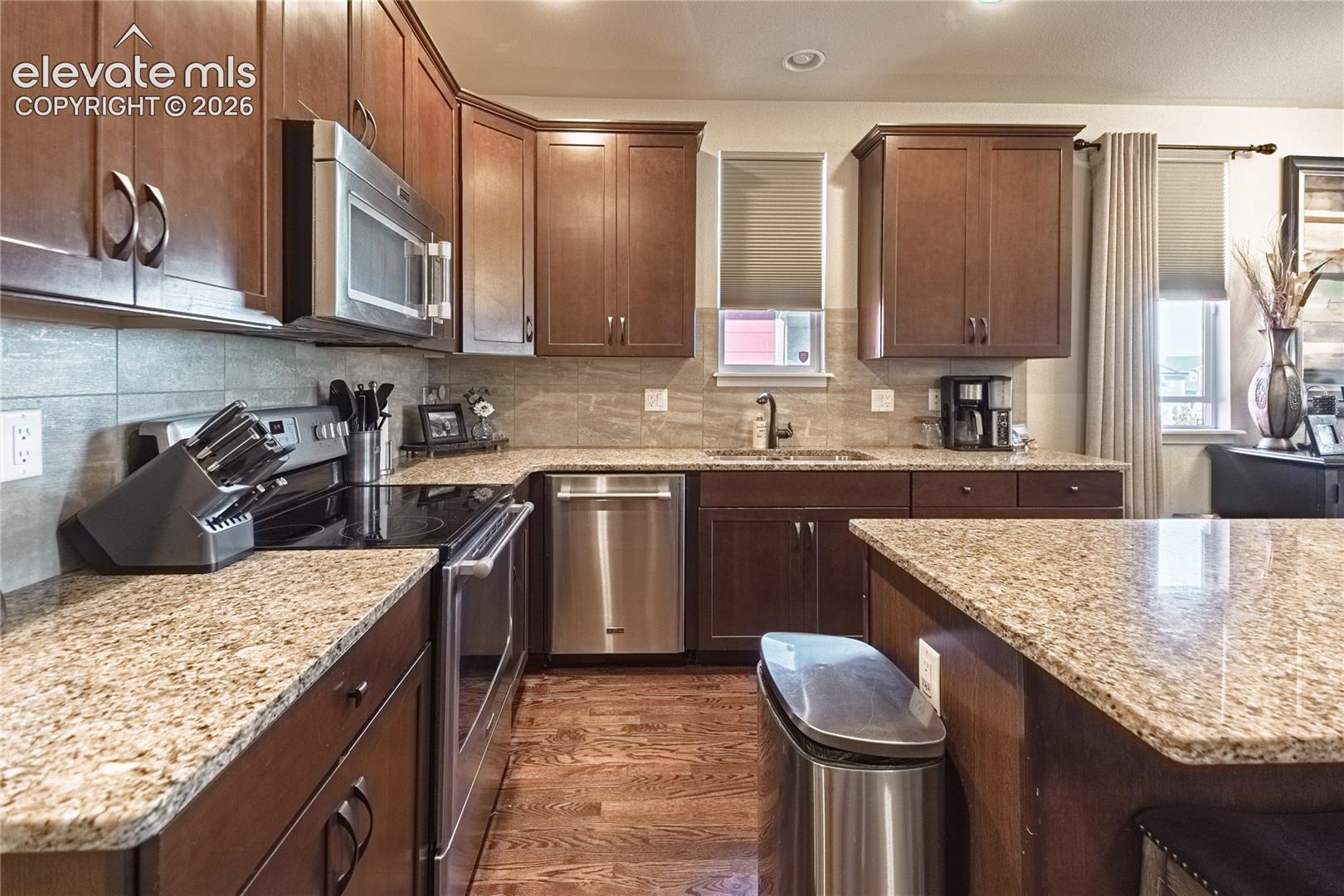 Kitchen featuring dark wood cabinetry, stone-finish countertops, tile backsplash, stainless steel appliances, and wood-finish flooring