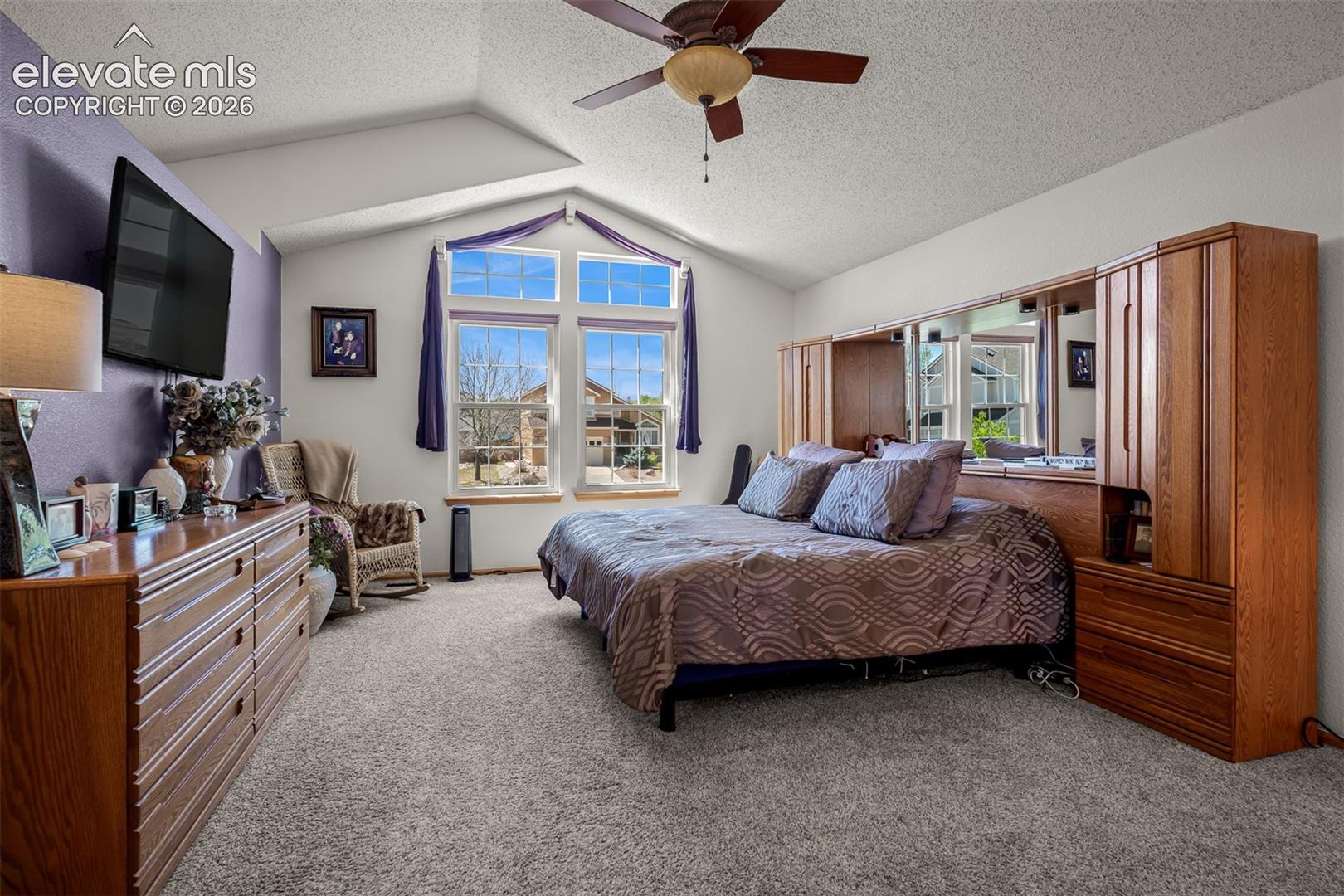 Vaulted ceilings enhance the large primary bedroom featuring a ceiling fan and a prominent window arrangement