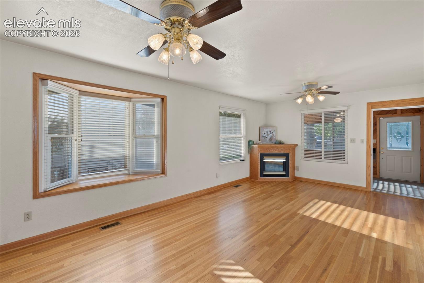 Hardwood flooring throughout, bay window with wood trim, ceiling fans with integrated lighting, and a corner fireplace