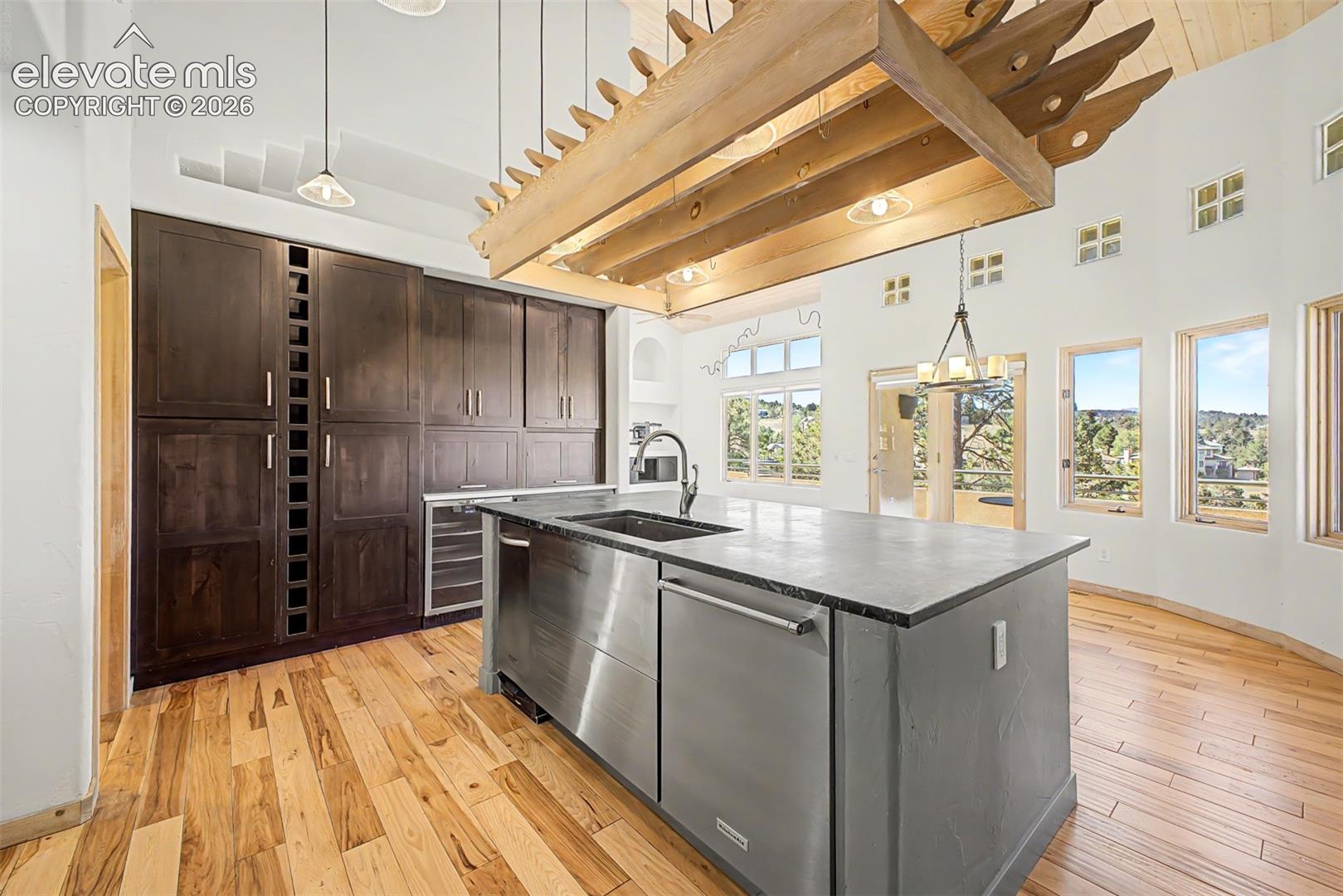 High ceilings with exposed wooden beams, dark wood cabinetry with integrated wine storage, stainless steel appliances, a center island with a sink, and light wood-finish flooring