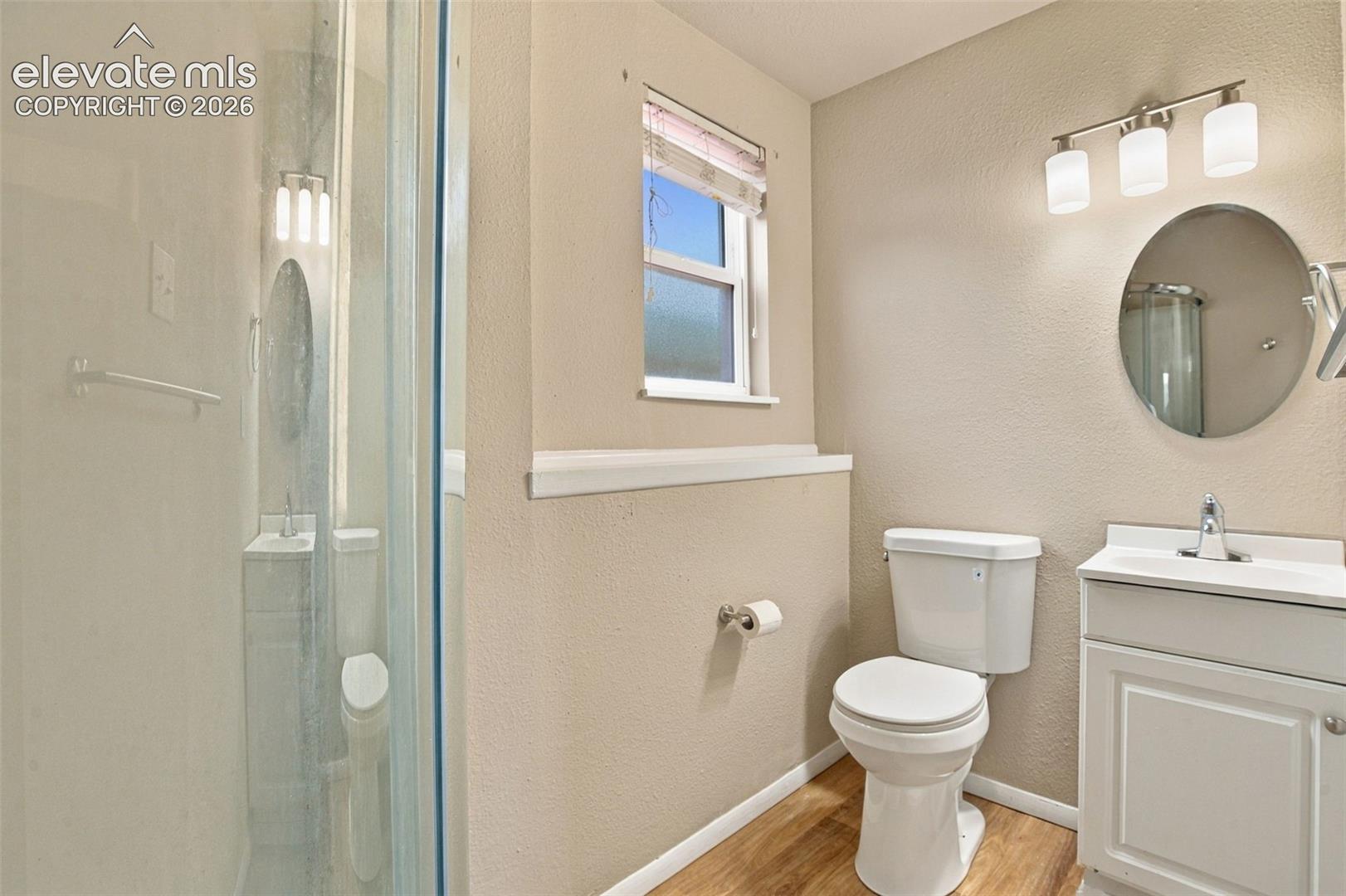 Bathroom featuring a glass-enclosed shower, white vanity cabinet, circular wall mirror, three-light fixture, and wood-finish flooring