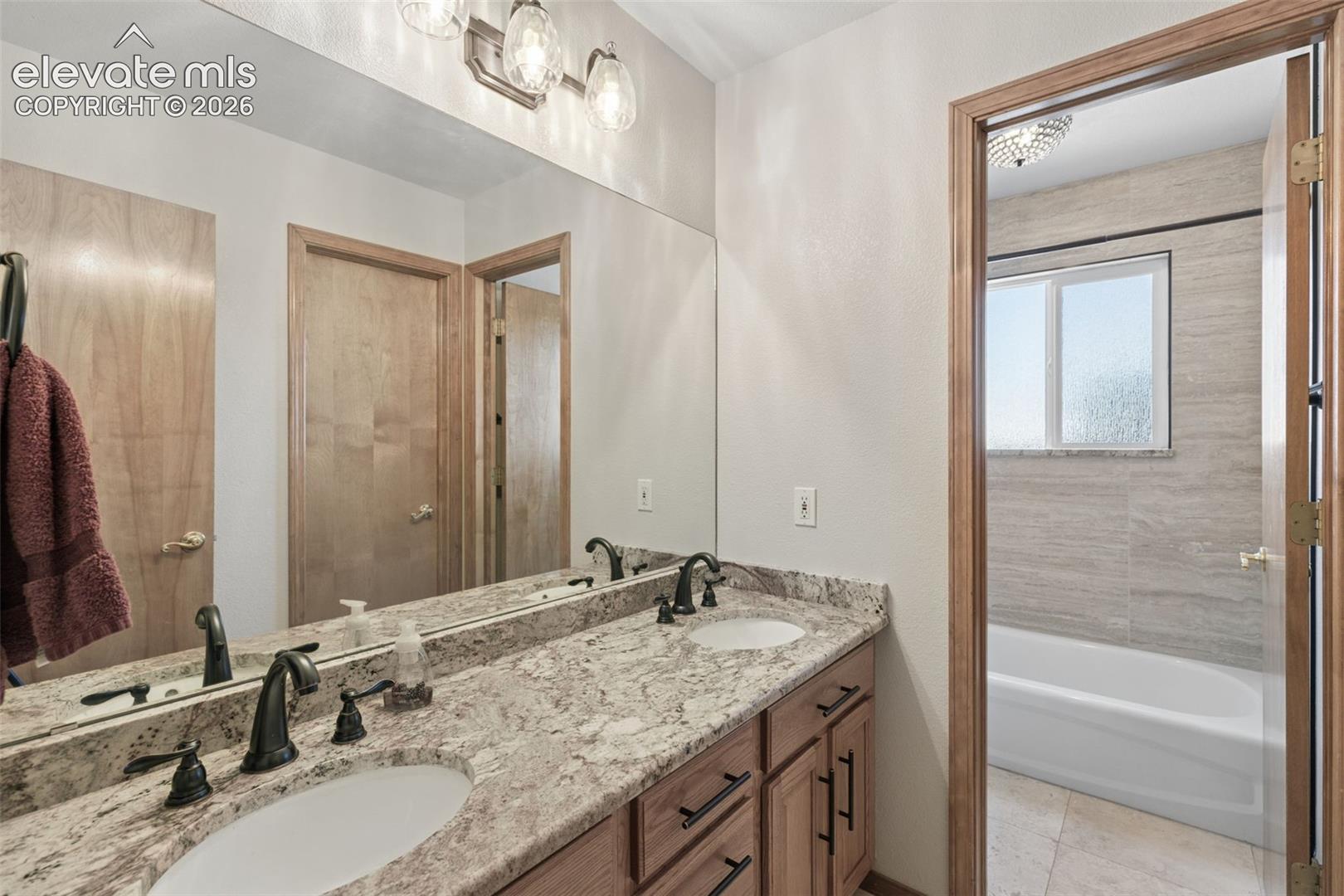 Dual-sink vanity with natural stone countertops, under-mount basins, dark hardware, and large-format wall mirror, including a separate bathing area with a tiled tub surround and window