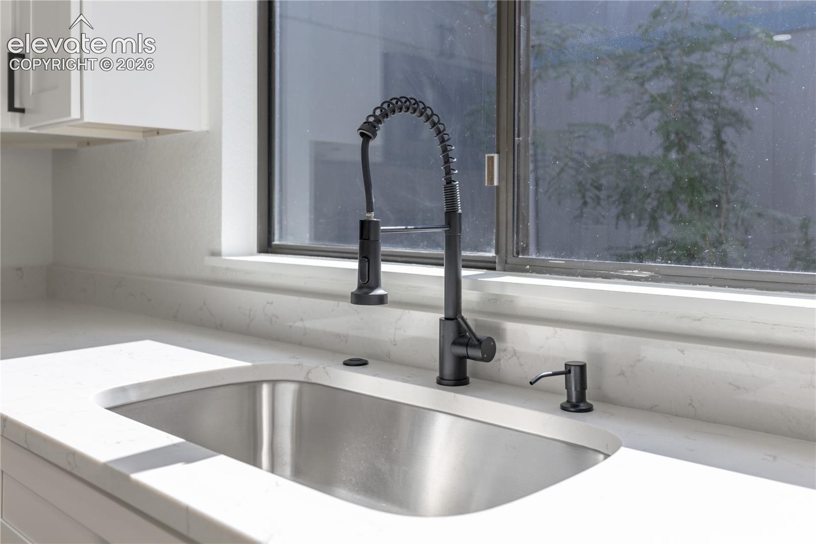 Undermount stainless steel kitchen sink featuring a matte black pull-down spring faucet, integrated soap dispenser, white stone-composite countertop, and white cabinetry