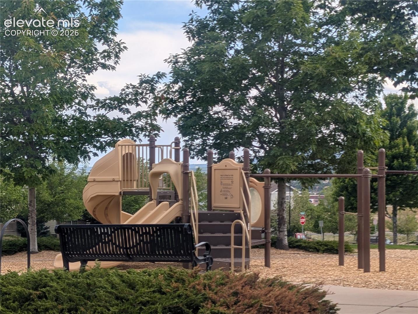 Outdoor playground featuring a tan slide structure, steps, and a black metal bench