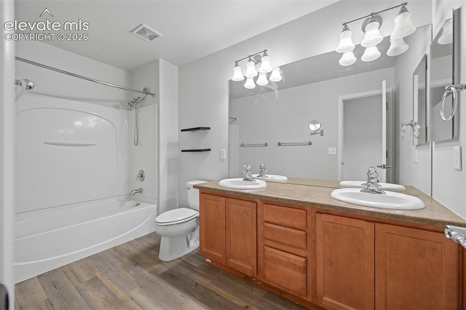 Bathroom featuring a dual vanity with wood-finish cabinetry and integrated sinks, a shower-tub combination with a white surround, wood-finish flooring, and wall-mounted lighting fixtures