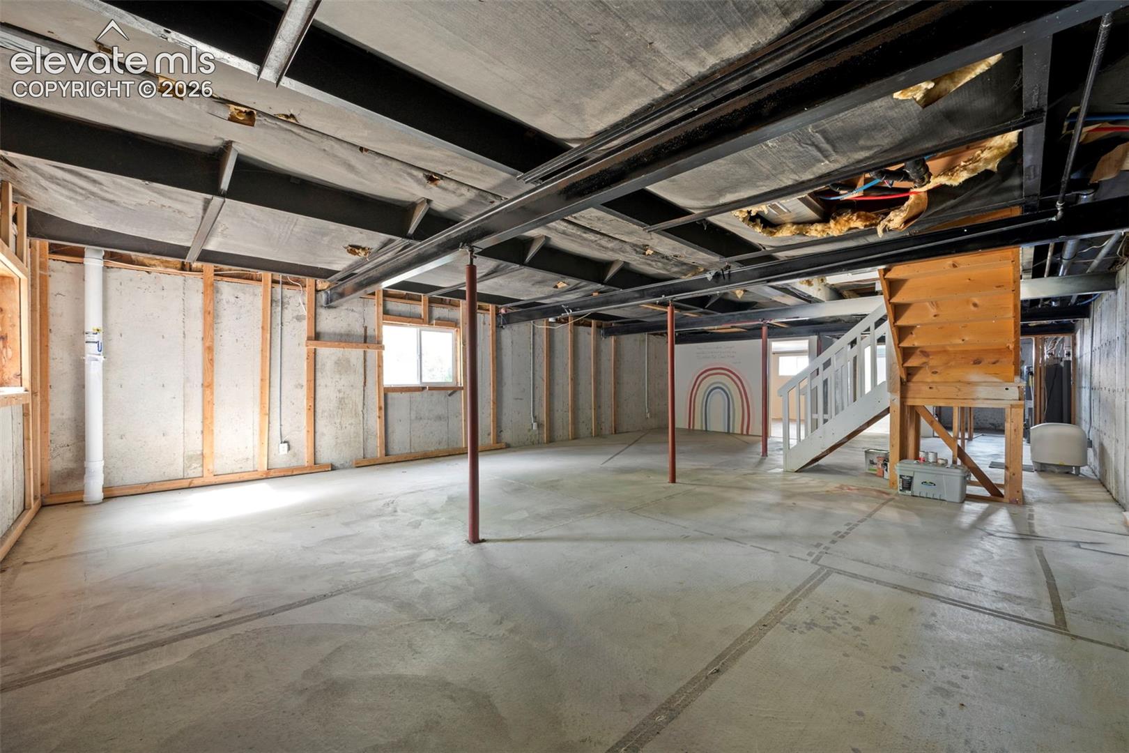 Unfinished basement space featuring exposed steel support beams, concrete flooring, wood wall framing, and a wooden staircase