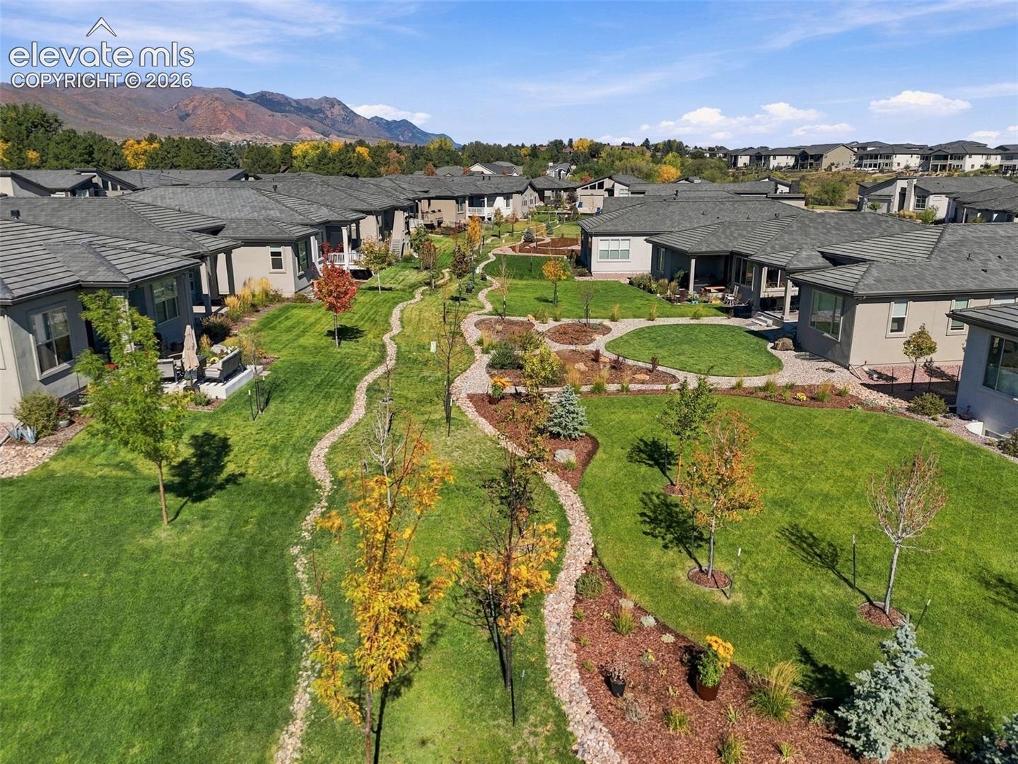 Community landscape featuring expansive turf lawns, winding stone pathways, curated garden beds, mature trees, and a mountain backdrop