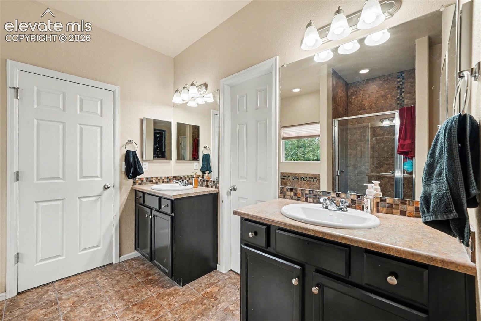 Primary bath with dual vanity sinks, dark wood-finish cabinetry, a full-width vanity mirror, and a mosaic tile backsplash