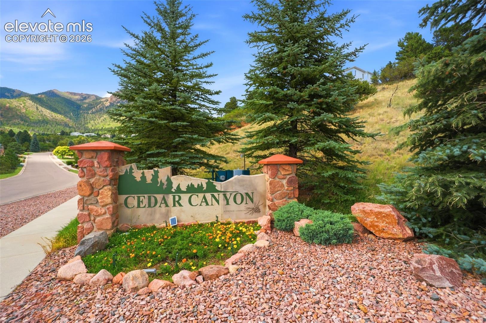 Stone-clad monument sign for Cedar Canyon featuring integrated landscaping, mature evergreen trees, and a mountain backdrop