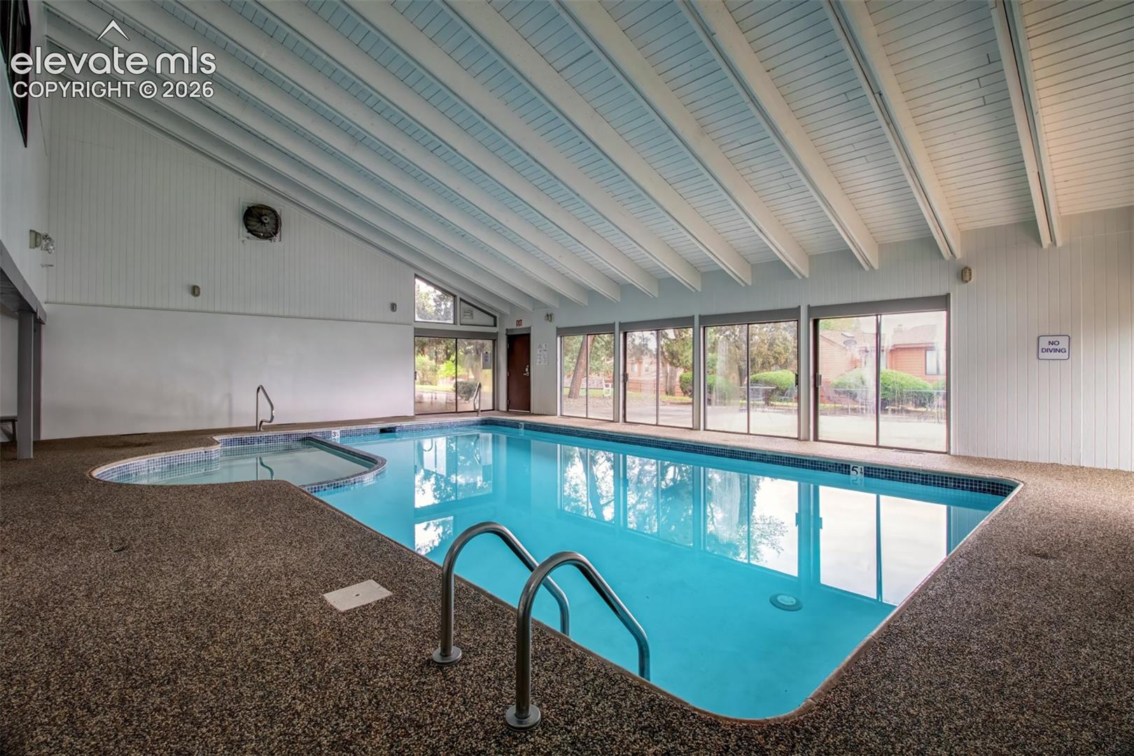 Indoor swimming pool with an integrated spa, featuring vaulted beamed ceilings, expansive glass sliding doors, and textured aggregate flooring
