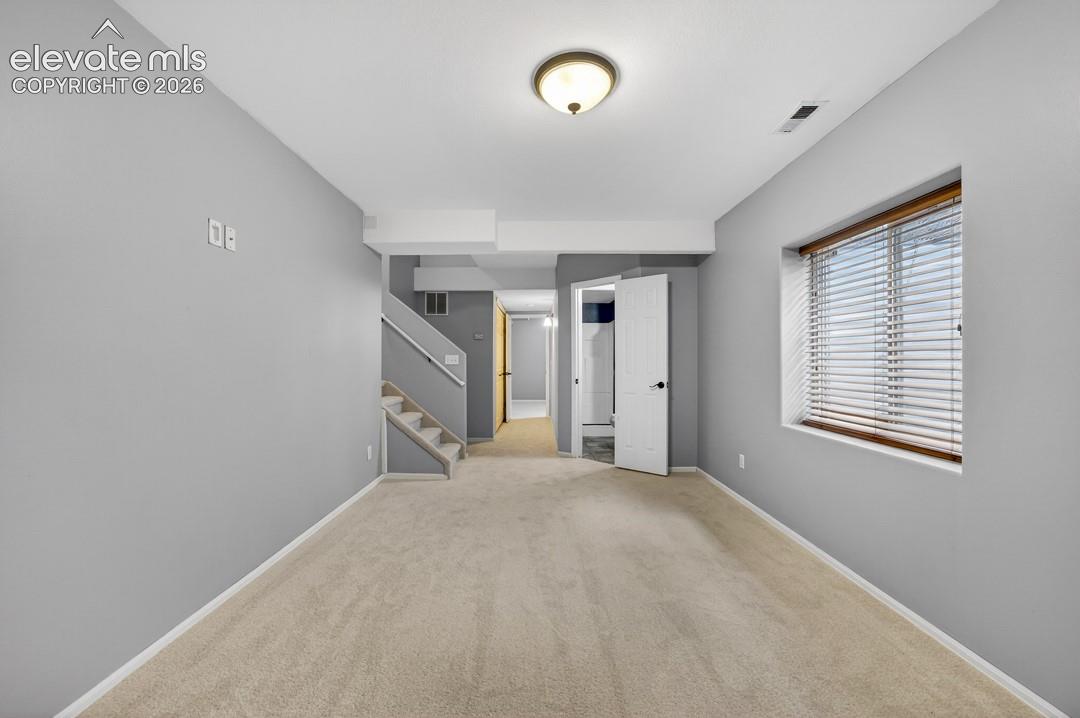 Neutral-toned interior featuring wall-to-wall carpeting, recessed window with horizontal blinds, flush-mount ceiling light fixture, and staircase with a metal handrail