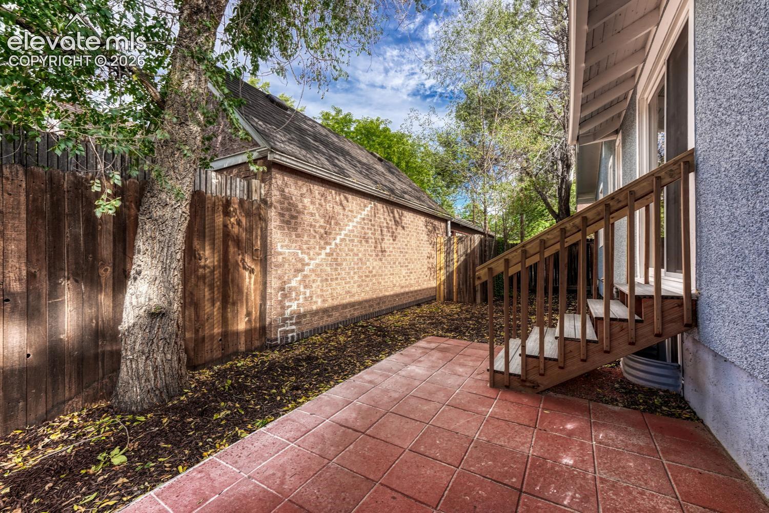 Patio area featuring red paver tiles, wooden privacy fencing, mature trees, and a brick-sided structure