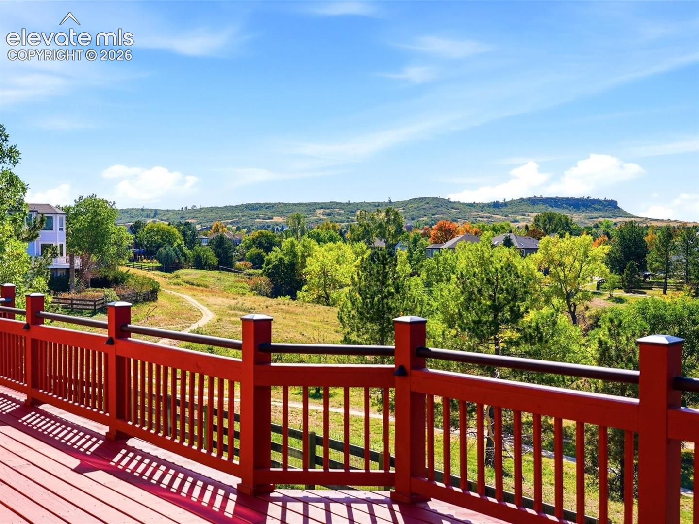 Elevated deck with a red-painted wood railing, featuring an expansive landscape of mature trees, grassy terrain, and a mountain backdrop