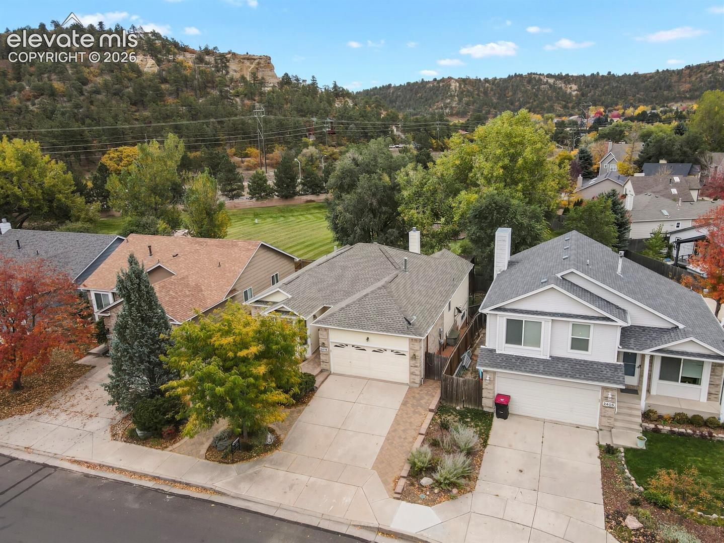 Aerial view featuring multi-level residential homes with shingle roofing, attached multi-car garages, paved driveways, and proximity to expansive natural mountain terrain