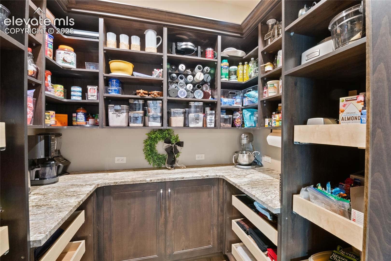Expansive walk-in pantry featuring granite counters, custom shelving, pull-out drawers, and dedicated prep space with secondary appliance area.