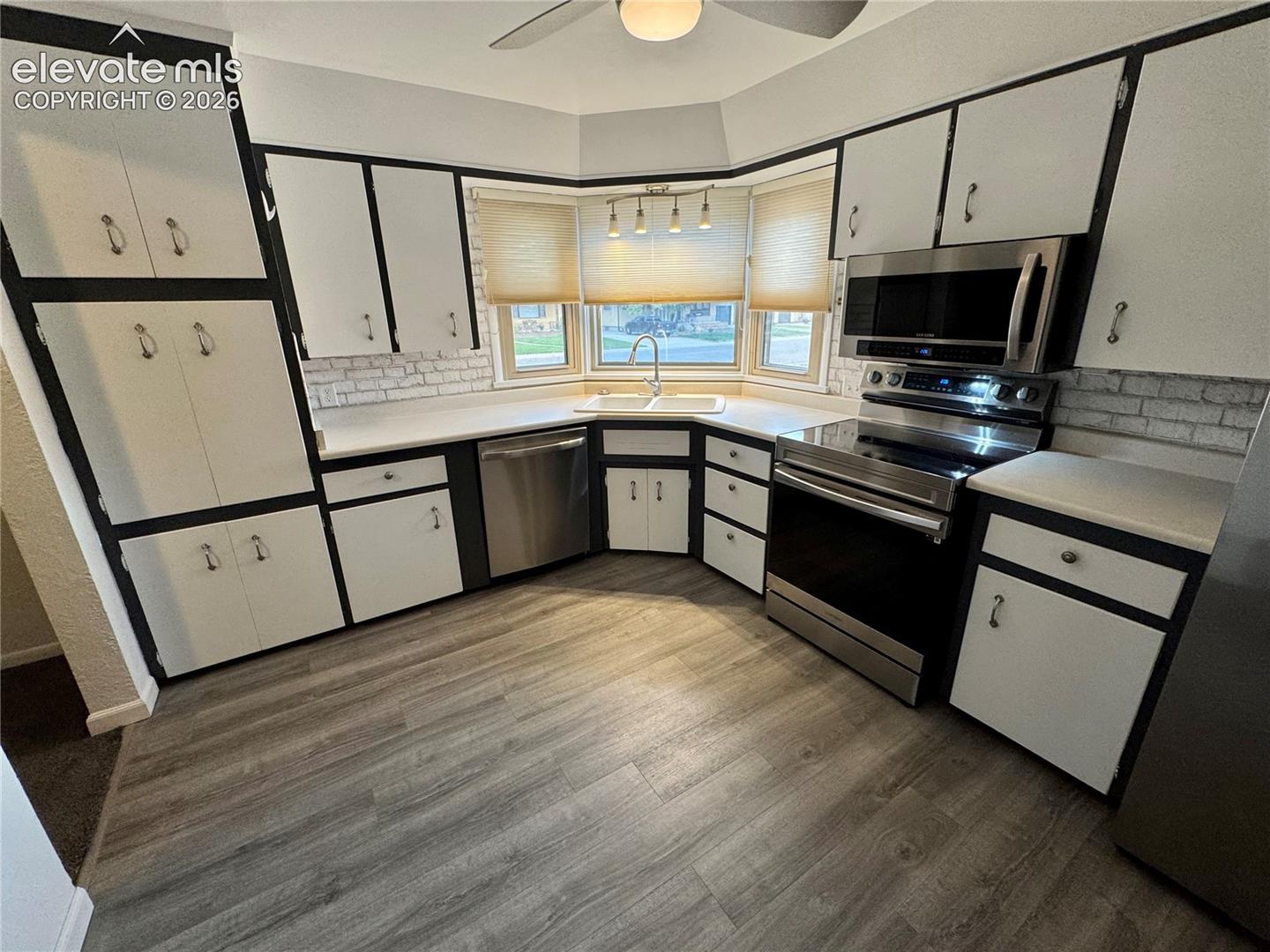 Kitchen featuring custom cabinetry with dark trim, stainless steel appliances, wood-finish flooring, a bay window, and a tile backsplash