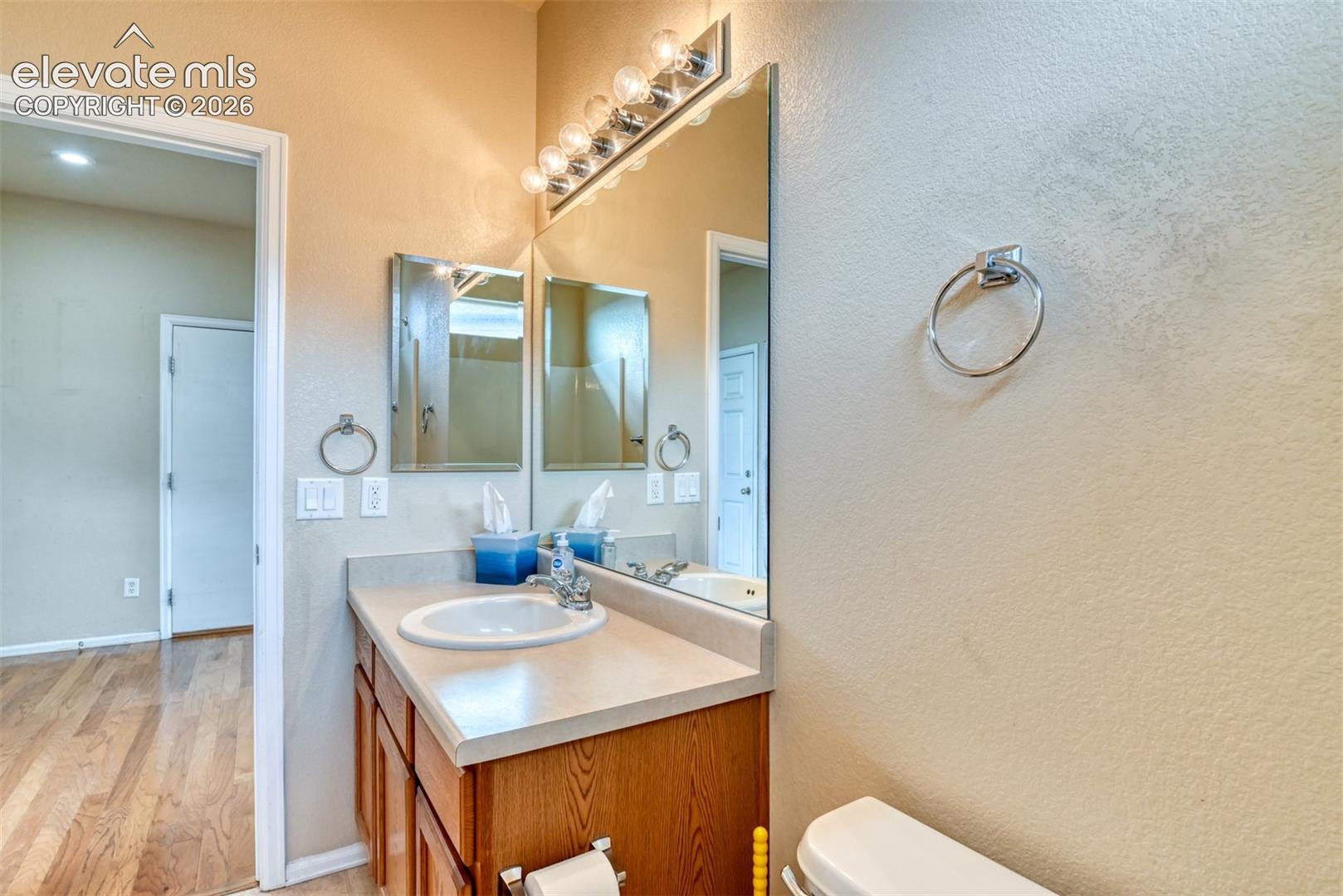 Bathroom featuring a wood-finish vanity, solid surface countertop with integrated sink, large wall-mounted mirror, multi-bulb overhead vanity light, and metal towel rings