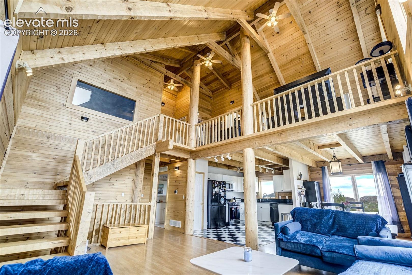 Expansive great room featuring a vaulted wood-finish ceiling with exposed beams, wood-finish flooring, and a prominent staircase leading to an upper-level balcony