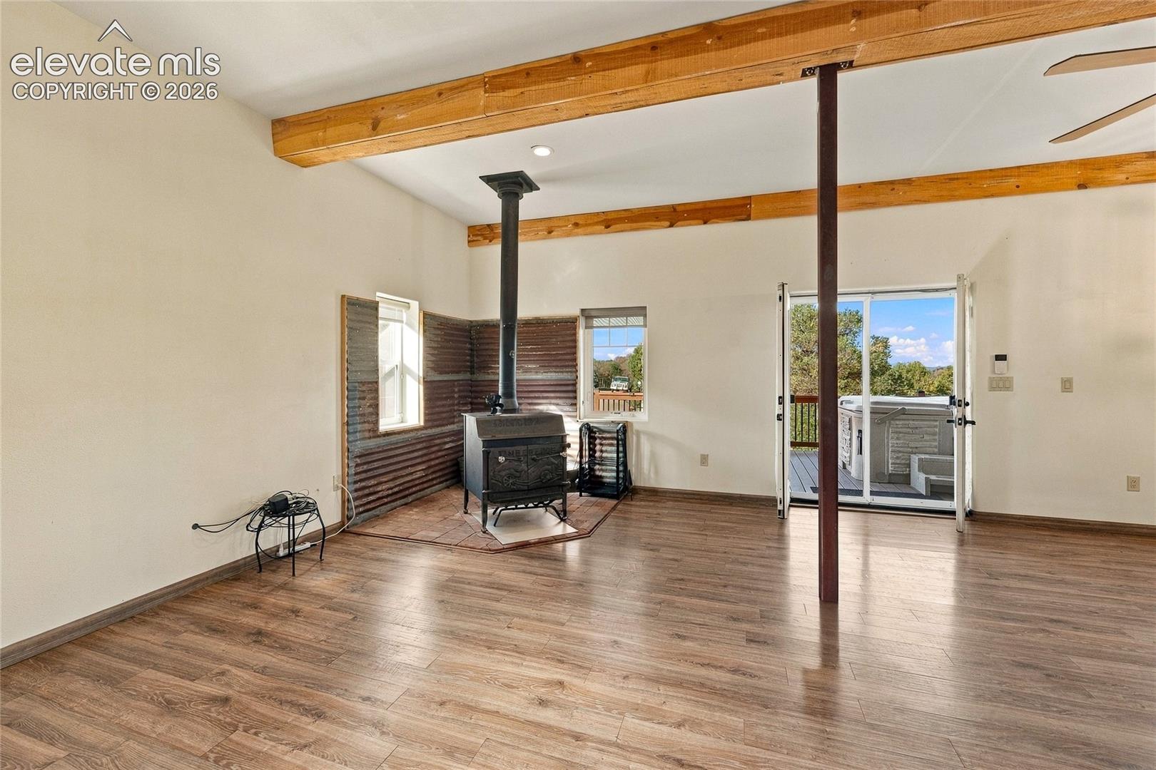 Open-concept interior featuring exposed wood ceiling beams, wood-finish flooring, a cast-iron wood-burning stove with corrugated metal accent wall