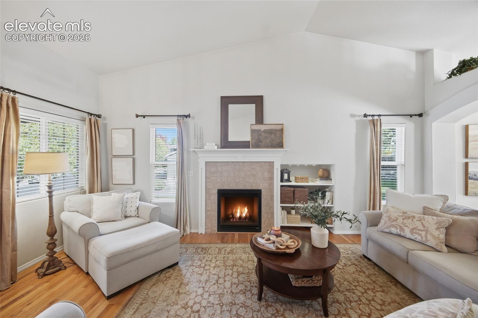 Vaulted ceilings, a tiled fireplace with mantel, built-in shelving, and wood-finish flooring