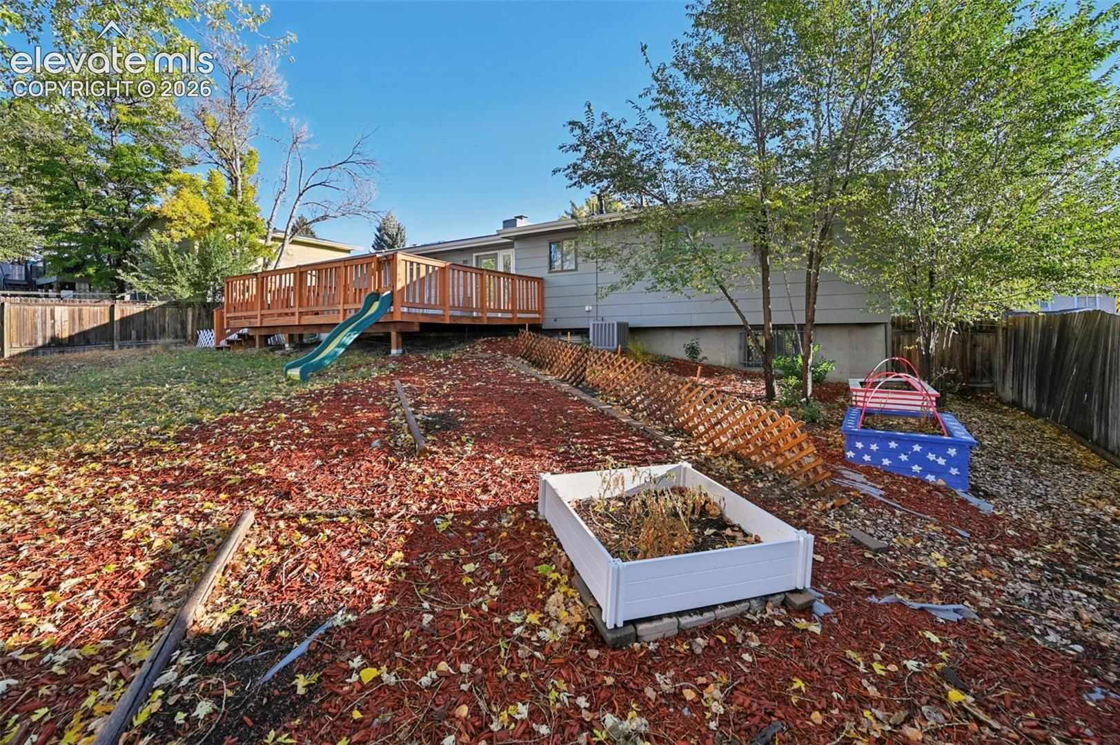 Backyard featuring a raised wooden deck with an attached sliding board, wood-slat perimeter fencing, and multiple raised garden beds