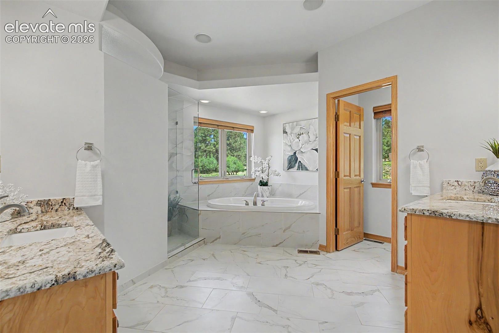 Bathroom featuring a freestanding soaking tub, a clear glass shower enclosure, dual vanity sinks with light-toned countertops, light wood-finish cabinetry, and white tiled flooring