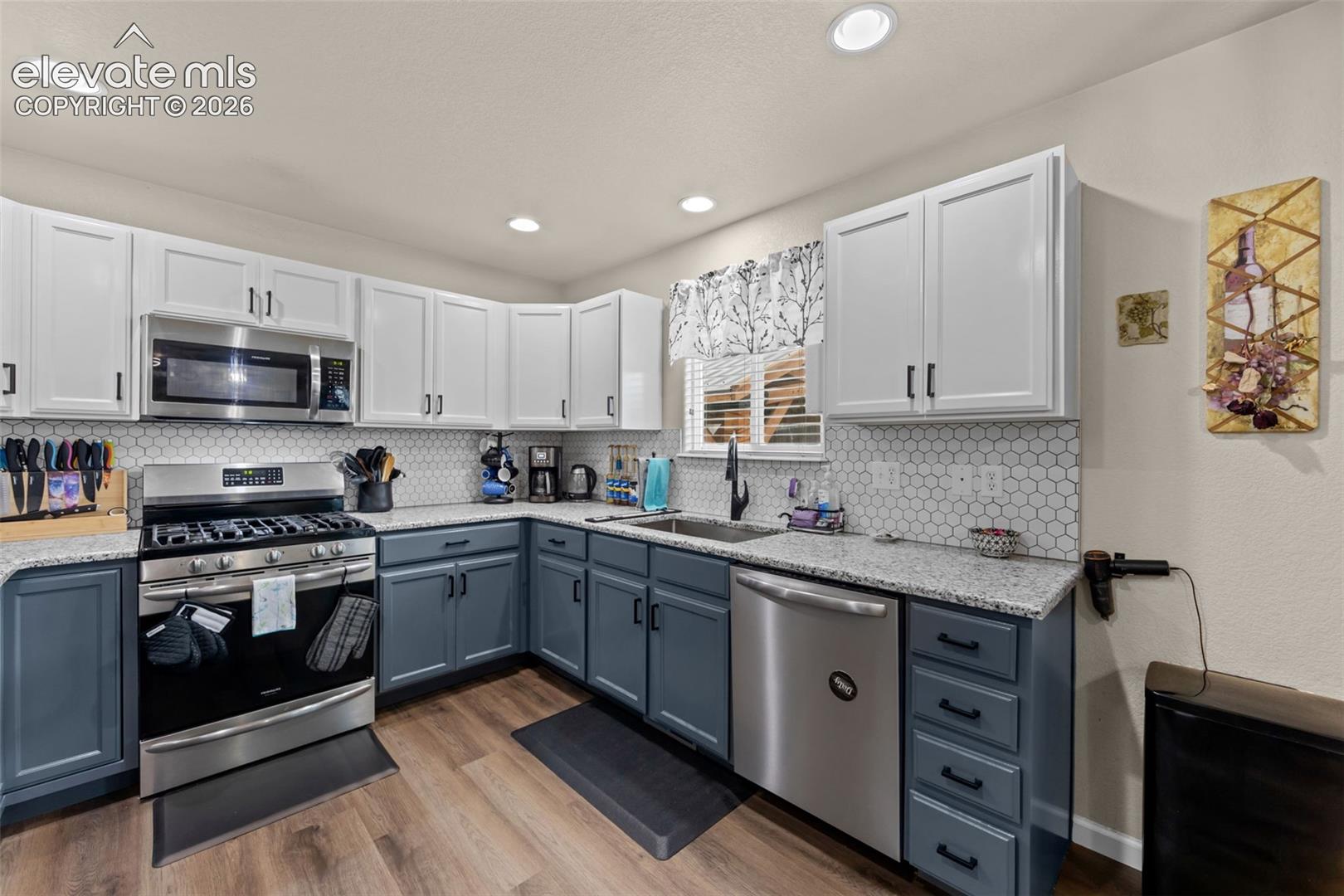 Two-tone cabinetry with contrasting upper and lower units, stainless steel appliance suite, hexagonal tile backsplash, stone-finish countertops, and wood-finish flooring
