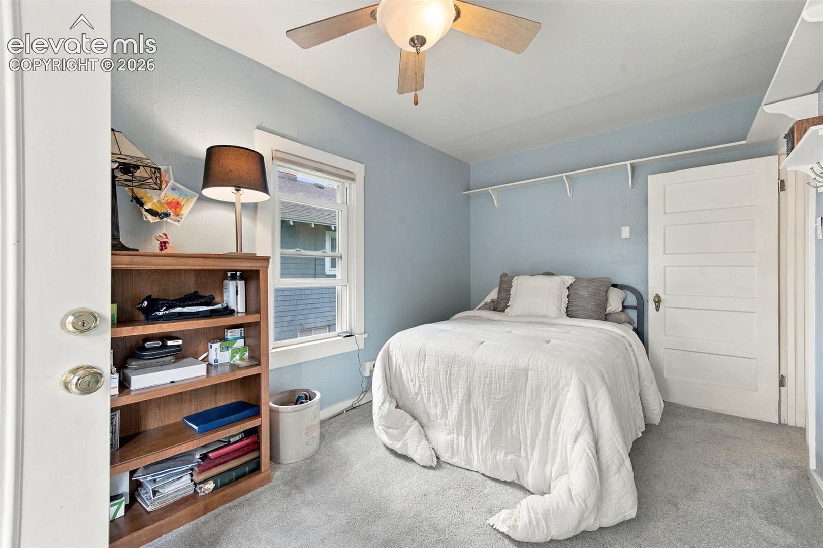 Neutral-toned carpeted room featuring a ceiling fan with integrated light fixture, a single-hung window with casing, a multi-panel interior door, and wall-mounted shelving