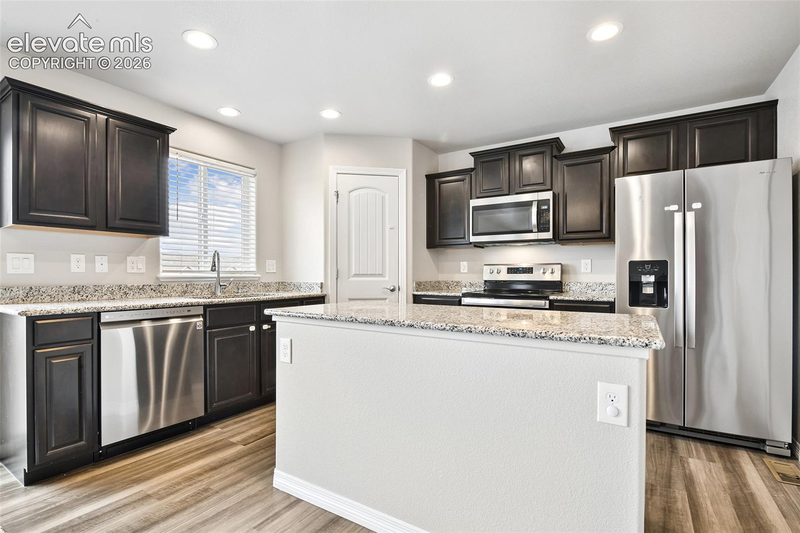 Kitchen featuring dark-finish cabinetry, stone-surfaced countertops, a center island, stainless steel appliances, and wood-finish flooring