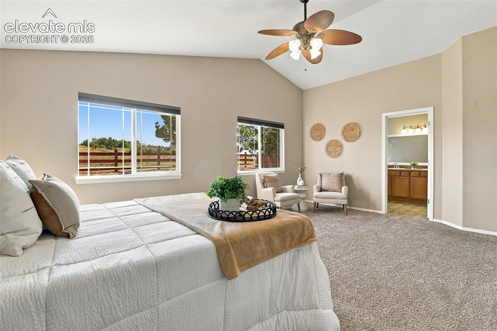 Vaulted ceilings, a ceiling fan with integrated lighting, expansive windows, carpeted flooring, and an open doorway to a bathroom with a vanity