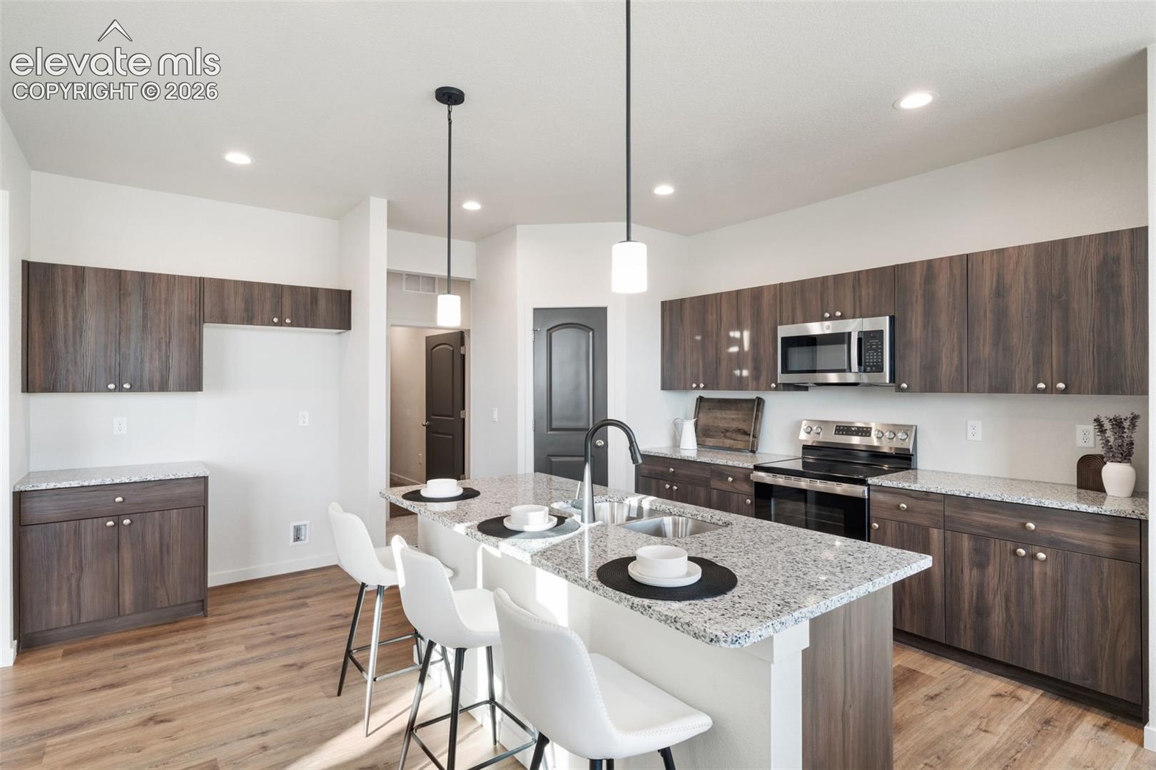Kitchen featuring wood-finish cabinetry, light-toned countertops, stainless steel appliances, a center island with a sink, and pendant lighting