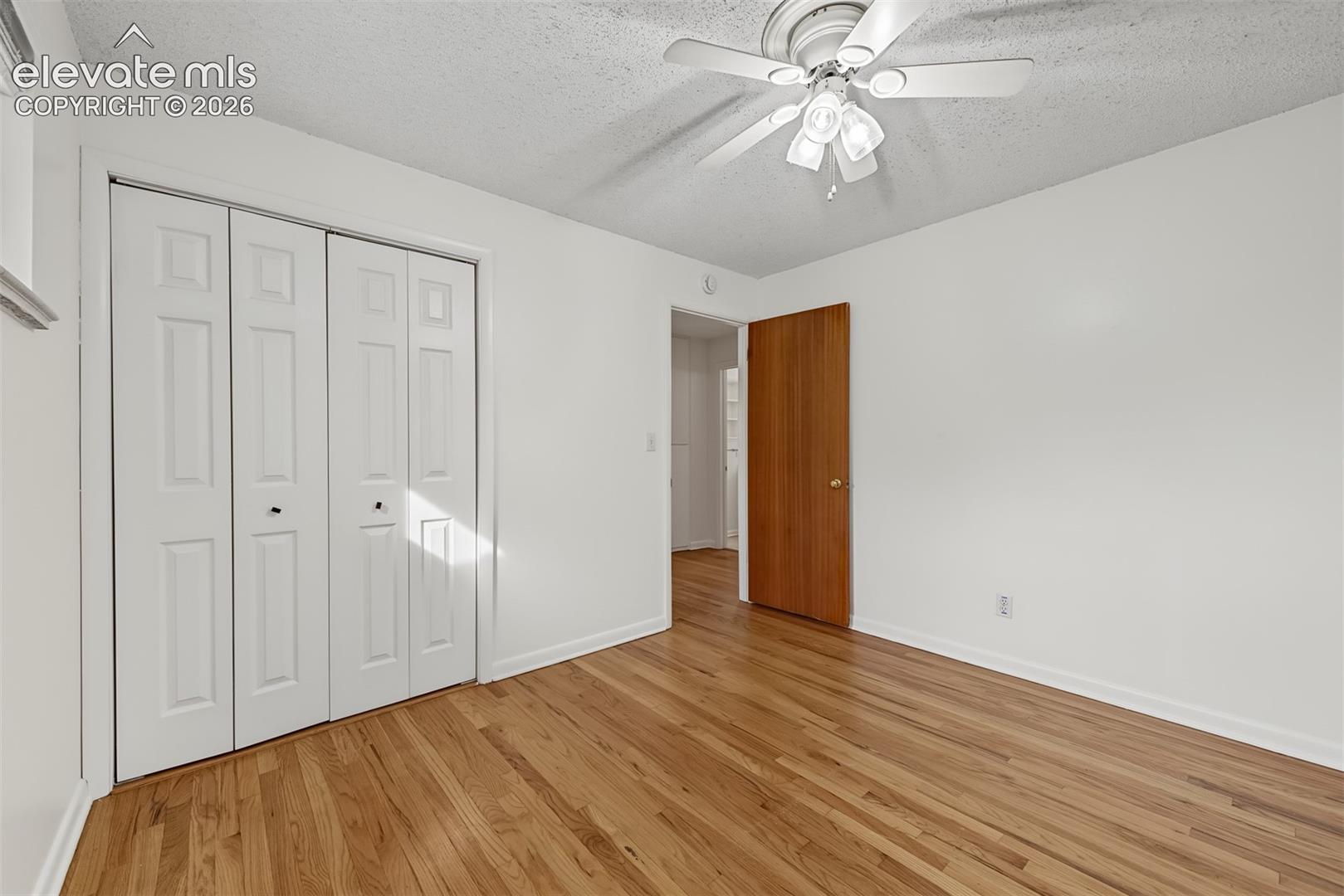 Room featuring wood-finish flooring, white four-panel bi-fold closet doors, a natural-finish wood door, and a multi-light ceiling fan