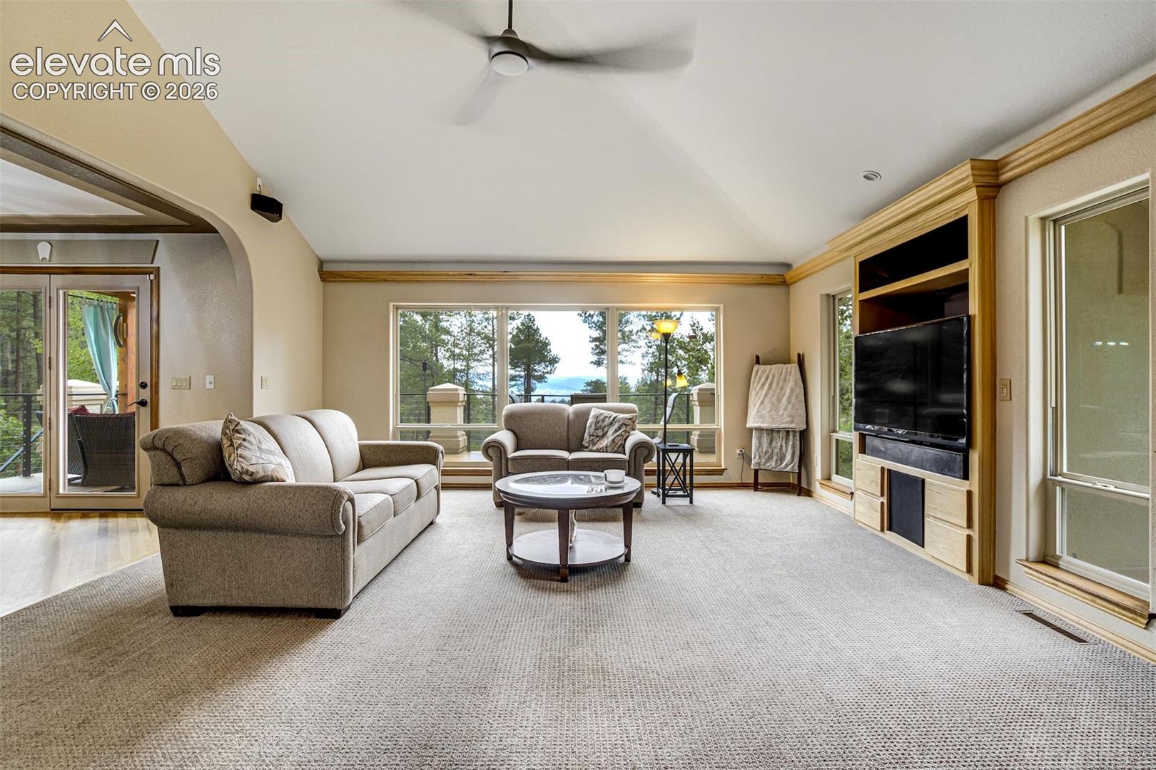 Spacious primary living area featuring expansive windows and glass sliding doors framing natural light, neutral wall finishes, engineered wood-finish flooring, a ceiling fan, built-in entertainment cabinetry with a mounted television, and a round cocktail table anchoring the seating arrangement