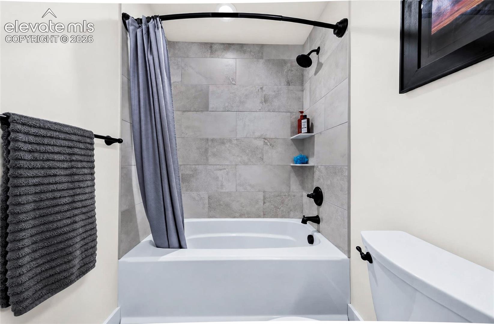 Bathtub surround featuring large-format light grey rectangular tiling, integrated corner shelving, and matte black plumbing fixtures