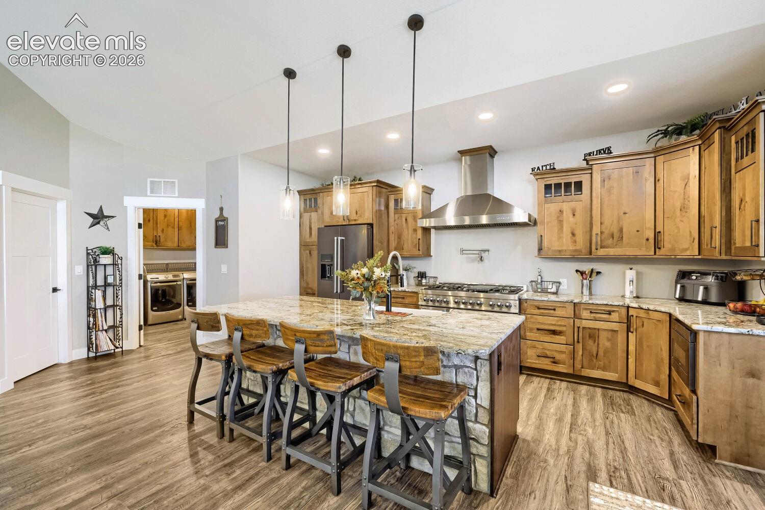 Kitchen featuring a stone-clad island with seating, granite countertops, stainless steel appliances, wood-finish flooring, and pendant lighting