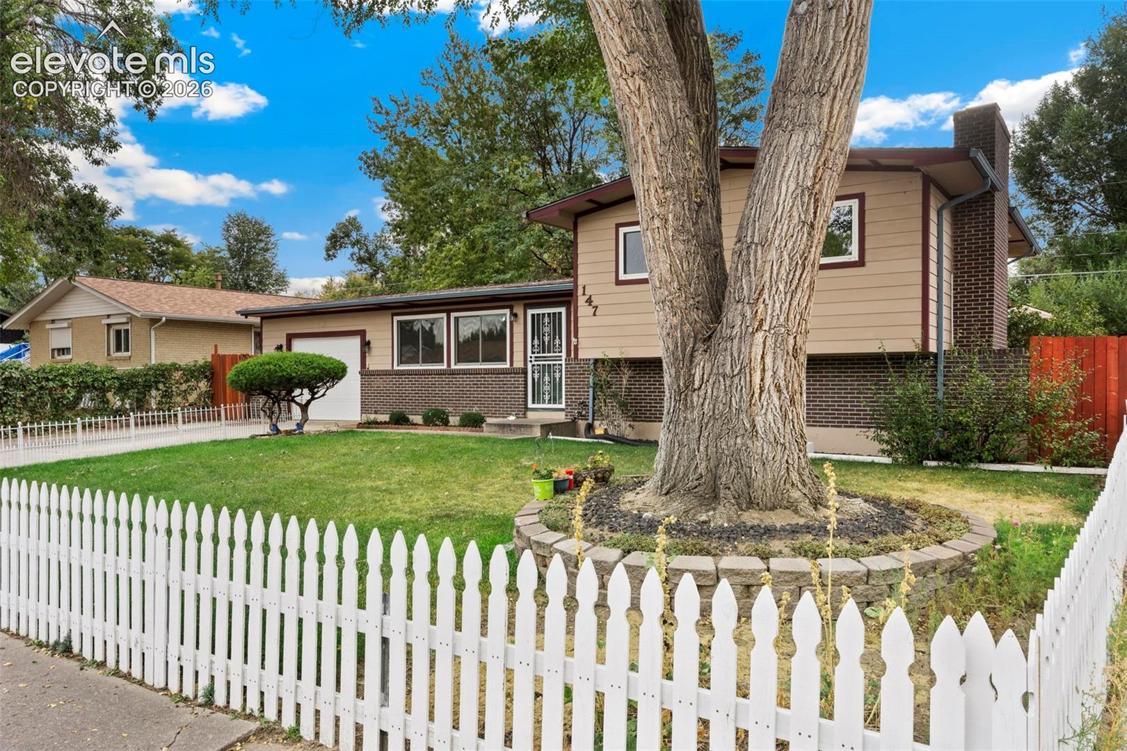 Picket fence front yard with a mature tree, manicured lawn, and an attached garage